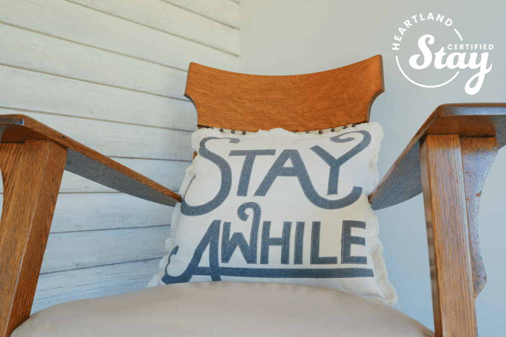 A padded wooden chair features a decorative pillow with the text "STAY AWHILE."