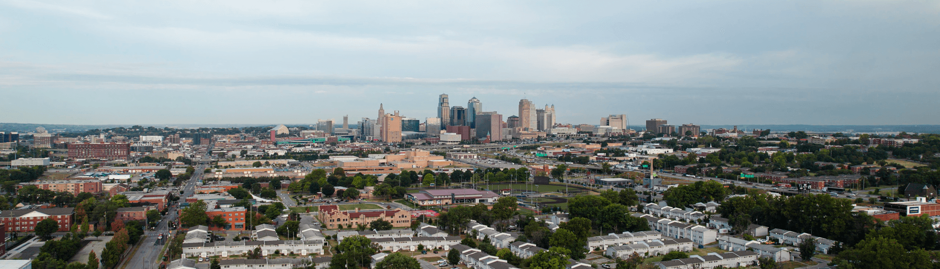 A panoramic view of a city skyline with various buildings and green spaces under a cloudy sky.