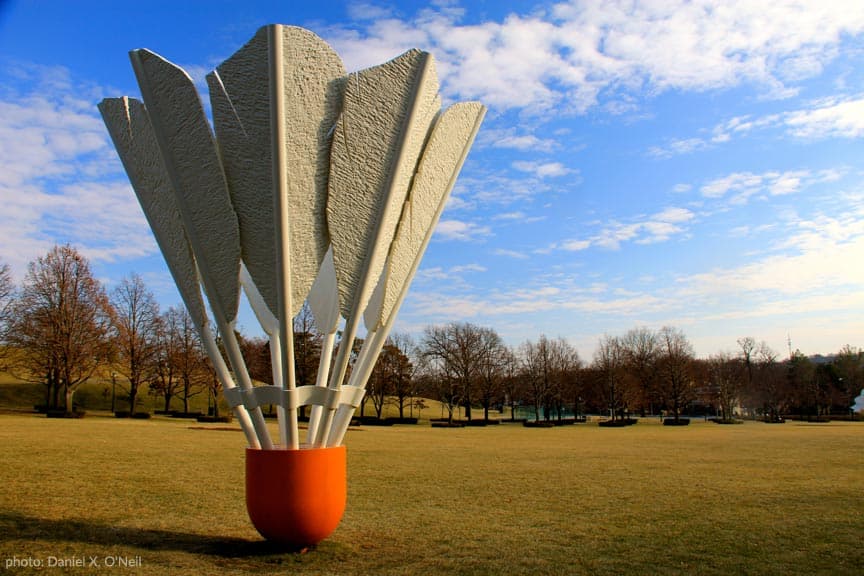 A large, abstract sculpture resembling a bouquet of petals stands on a grassy field under a blue sky.