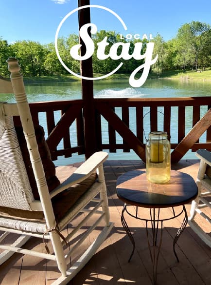 A peaceful lakeside porch featuring rocking chairs and a small table with a jar, framed by greenery.