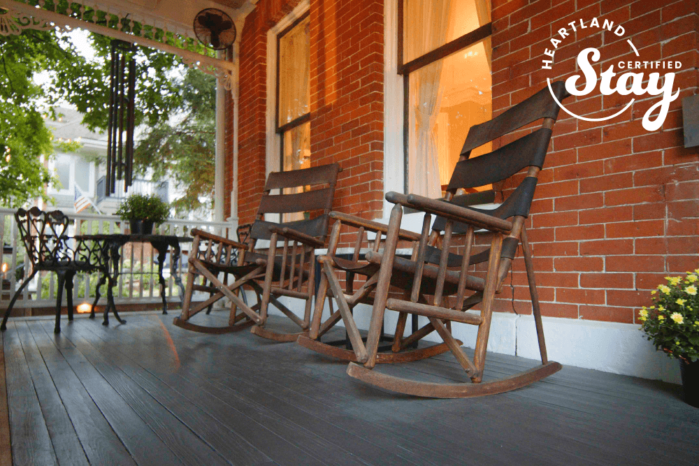 A cozy porch featuring wooden rocking chairs and a small table, surrounded by greenery.