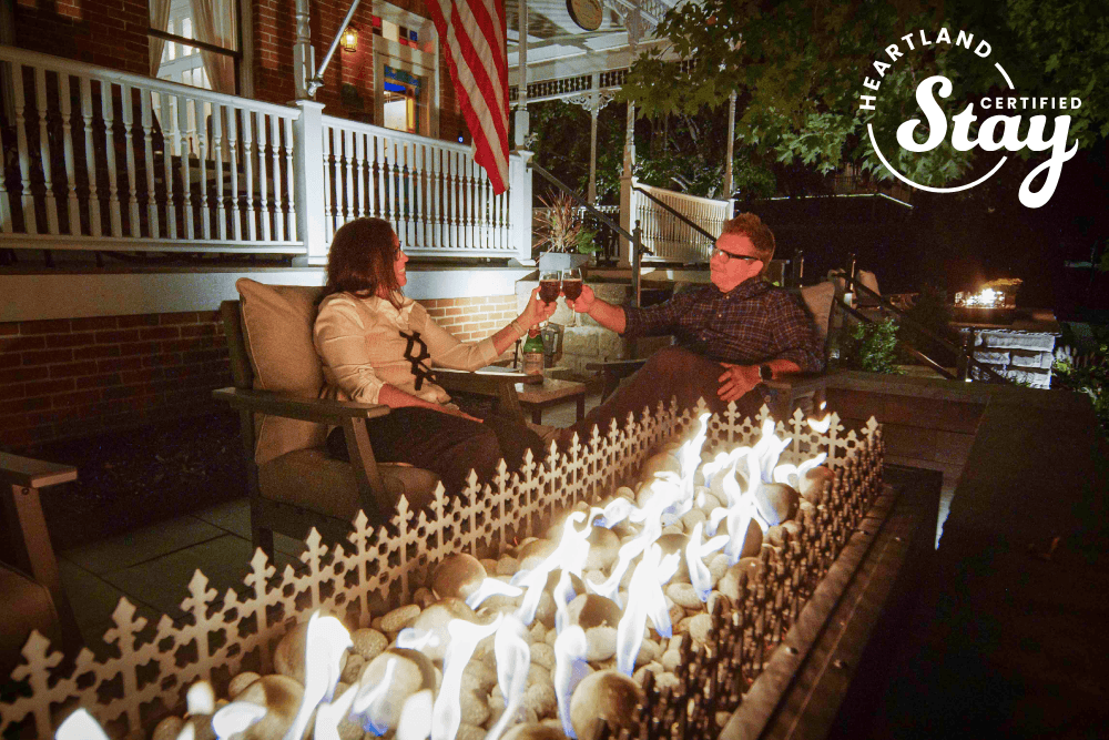 A couple clinks glasses by a cozy fire pit on a porch at night, with an American flag in the background.