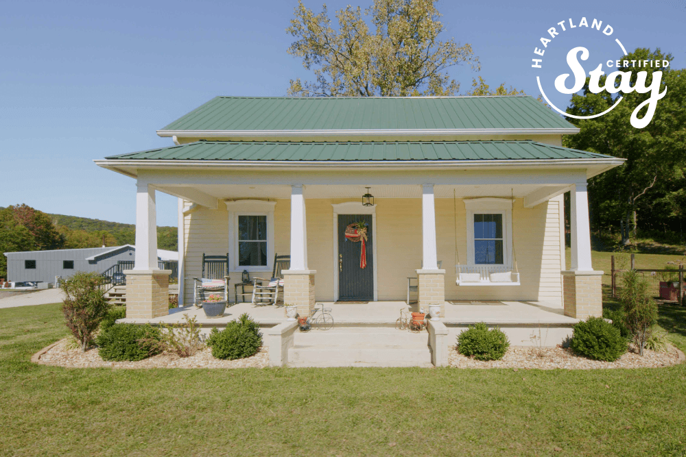 A charming yellow house with a green metal roof and front porch surrounded by greenery.